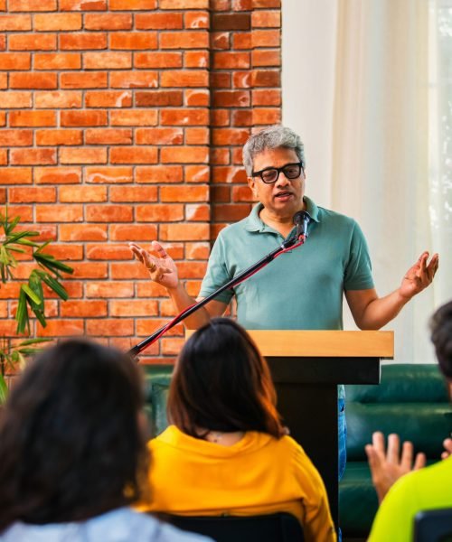 Indian Asian life coach, professional mentor delivering inspiring speech on business growth entrepreneurship to group of young startup founders at conference, speaking confidently on podium with a mic